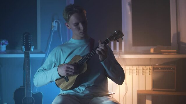 An adult smiling man plays the ukulele in the light neon dark room. A strong and blond guy sits and plays a small guitar in atmosphere place. A happy person plays a musical instrument.