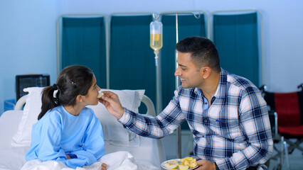 A caring Indian father is feeding fruits / apples to his little daughter - a nutritious meal  a healthy diet  juicy. A handsome Indian guy with his sick little girl-child in the hospital - father-d...