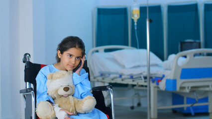 A sweet little girl is talking to her family members on mobile in the hospital - loneliness an internet connection. A cute little kid wearing blue hospital clothes is sitting in a wheelchair with ...