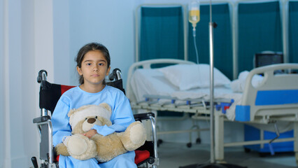 A little child patient is sitting in a wheelchair while holding a teddy bear - a favorite toy an accident an injury. A little kid is alone in the hospital ward - a recovery room a medical clinic...