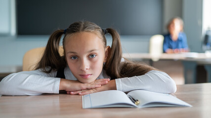 Little caucasian girl is bored at the lesson at school. The schoolgirl folded her head on the desk and the teacher sits in the background. 