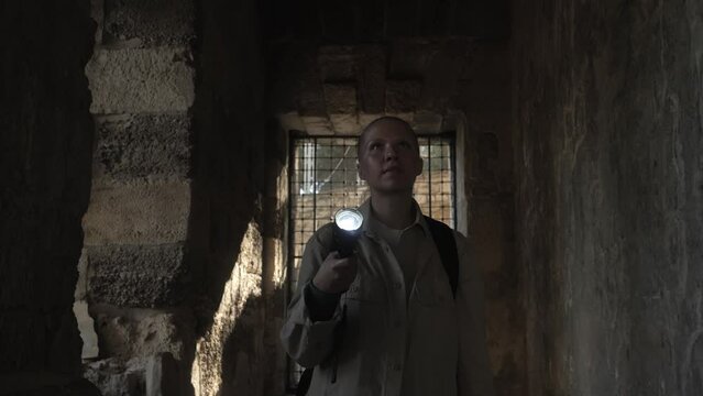 female archaeologist or student moves along a dark ancient corridor with a lantern in her hands and shines on the walls and ceiling around her with an anxious or inquisitive expression on her face. 4K