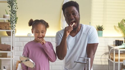 Black man with little daughter brushing their teeth together in bathroom during morning routine
