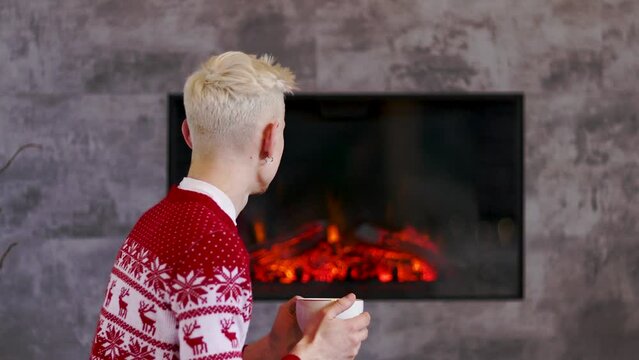 Young successful handsome man in a Christmas sweater resting with a cup of tea by the fireplace