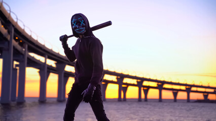 Street hooligan in black hoodie and mask with baseball bat at sunset near bridge