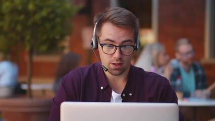 Attractive young man in headset having a video call on laptop sitting in the cafe. Realtime