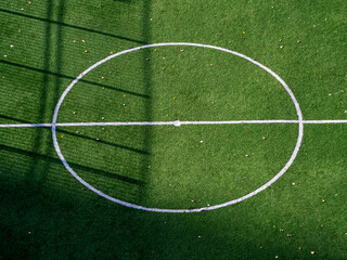The central circle on the green grass of the football field. View from above. Autumn leaves lie on the playing field, the shadow of the fence is visible.
