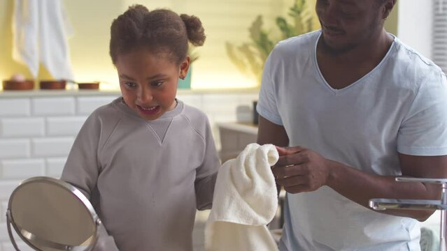 African American man helping his little daughter to wipe her face with towel after washing, soing morning routine together in bathroom