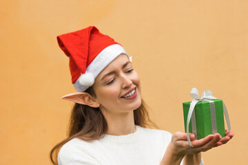Young smiling woman in santa hat with elf ears holding green gift box in front of her. Christmas...