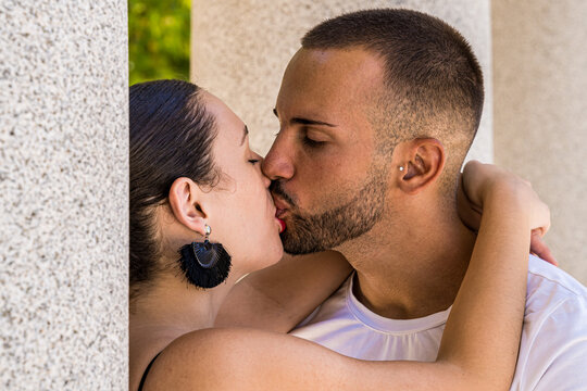 Close-up shot of a young couple kissing between stone columns, wearing black earrings and white shirt.