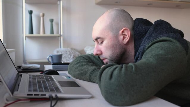 Young Caucasian bearded bald man is bored at his desk near laptop. Depressed person. Psychology and mental health.