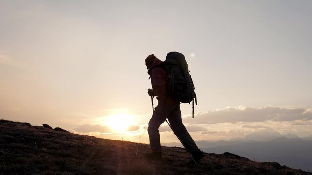 Man climber with backpack and trekking sticks poles raises up to top of cliff mountain. Walking at sunrise or sunset, sun rays shine background. Dressed in warm trekking clothes hiking. Shot in motion