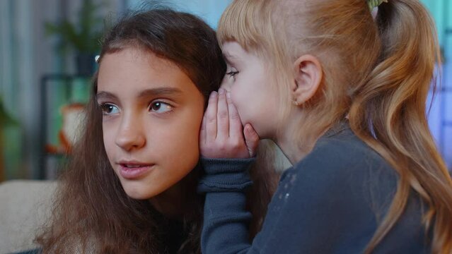 Teenage child whisper news rumors in ear to little sister kid girl share telling secrets gossip having fun. Female siblings children or best friends at home play room. Friendship, people communication
