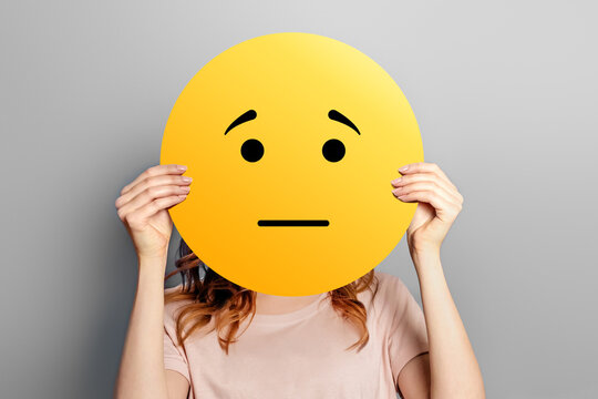 offended emoticon. Girl holds a yellow smiley with sad face isolated on a gray background