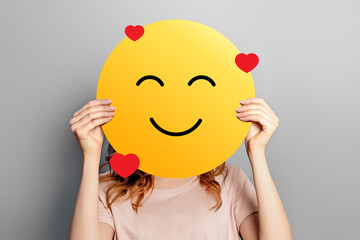 Emoticon in love. Girl holds a yellow smiley with romantic love face emoticon isolated on a grey...