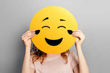 Laughing emoticon with tears of joy. Girl holds a yellow smiley with romantic love face emoticon isolated on a grey background
