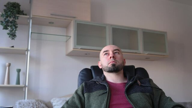Young Caucasian bearded bald man sitting on chear and thinking. Depressed person in living room. Psychology and mental health.