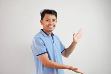 smiling asian man open arms pointing to the sides wearing blue t shirt isolated on white background