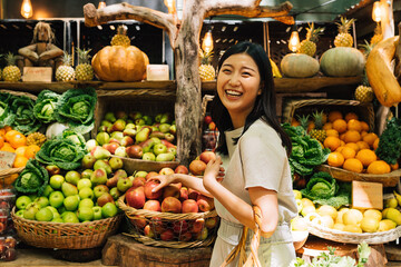 Laughing Asian woman on an outdoor market. Smiling female buying organic food. - obrazy, fototapety, plakaty
