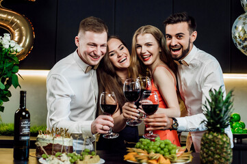 Friends at the festive table congratulate the birthday girl;
alcohol and snacks are on the table;
glasses with red wine in hands;
happy and joyful smiles;