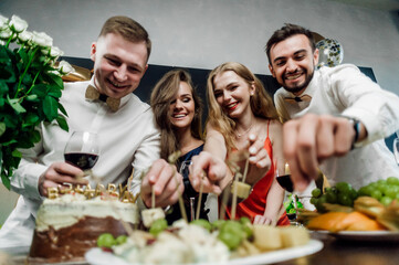 Friends at the festive table congratulate the birthday girl;
alcohol and snacks are on the table;
glasses with red wine in hands;
drink alcohol and socialize; are treated to snacks