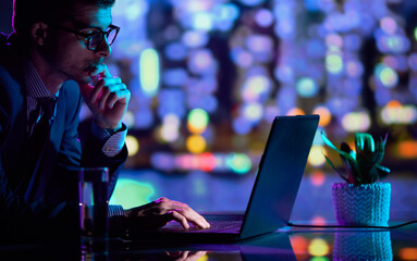Serious young man, office manager and IT specialist sitting at office and using laptop at evening. Cool colored neon hue lights in office space.