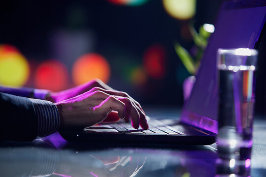 Cropped image of male hands of business man typing on laptop keyboard at evening time. Cool colored neon hue lights in office space.