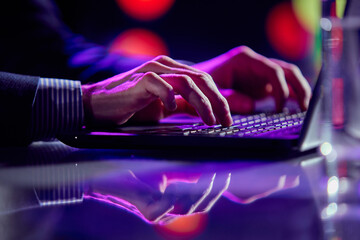 Cropped image of male hands of business man typing on laptop keyboard at evening time. Cool colored neon hue lights in office space. © Lustre Art Group 