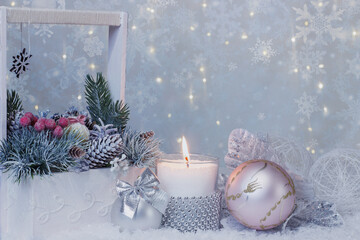 A white wooden basket with vintage lace and a bouquet of fir branches and cones, a burning candle in a decorative candlestick and two Christmas balls on a light blurred background of snowflakes 