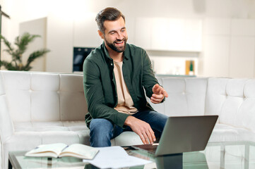 Obraz premium Online communication. Positive caucasian man, freelancer, sits on a sofa in living room, uses his laptop, talking online by video conference with colleagues or clients, gesturing hands, smiles happily