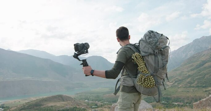 a happily smiling attractive man, a tourist, with a backpack on his back, records a video on the camera. he tells about his magnificent and interesting journey and points to the mountain with his hand