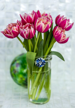 Pink tulips arranged in a glass vase with blue and white ceramic button on the rim placed on a counter top with a green glass ball in behind; Surrey, British Columbia, Canada