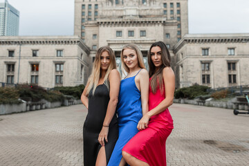 Three beautiful young happy women friends in a fashion elegant strappy evening dress stands in the city