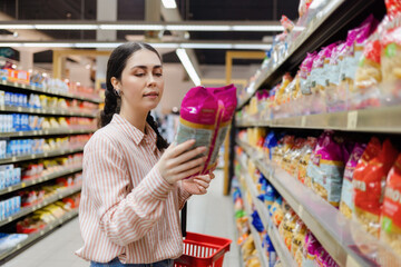 Shopping in grocery store. Portrait of young Caucasian woman choosing groceries in supermarket. Millennial is considering package of food. In background, rows of shelves with products