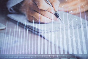 A woman hands writing information about stock market in notepad. Forex chart holograms in front....