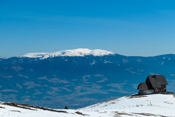 Panoramic view of mountain hut Wolfsbergerhuette (Wolfsberger Huette) on Saualpe, Lavanttal Alps, Carinthia, Austria, Europe. Looking at snow covered Koralpe. Alpine road leading to remote cottage