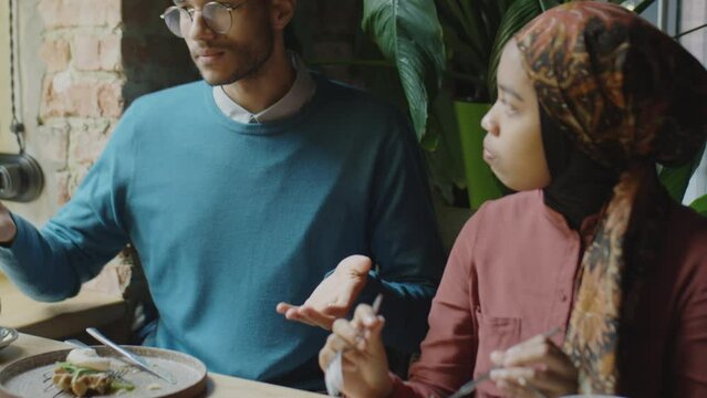 Over the shoulder shot of young man talking to Muslim woman and Afrcan American male colleague while eating lunch together in cafe