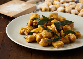 Plate of homemade gnocchi with sage butter sauce, garnished with sage leaves on a wooden table, with raw gnocchi in the background.