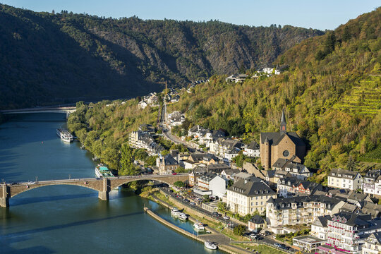 High view of riverside village with stone church and steep hillside slopes with stone bridge crossing; Cochem, Germany
