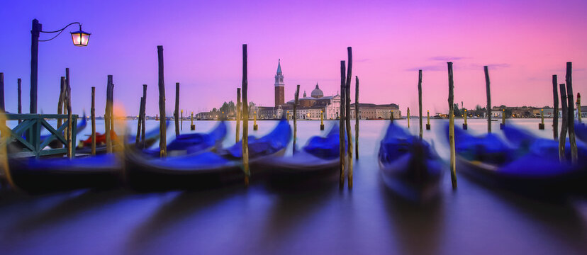 Gondolas moored along the shoreline of the Grand Canal during a vibrant sunset with a view of San Giorgio Maggiore and church in the distance; Venice, Italy