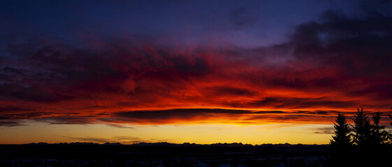 Dramatic colourful sky/clouds at sunset with silhouette trees and mountain range in background; Calgary, Alberta, Canada