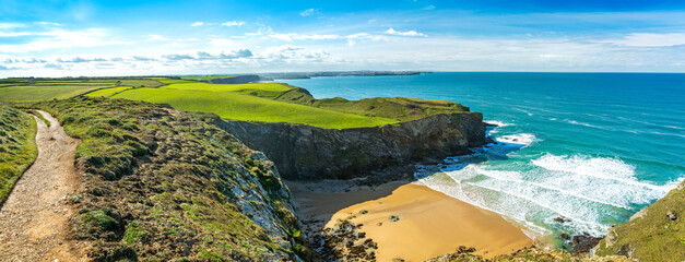 Gravel footpath on top of rocky cliffs on the shoreline with beaches and surf below with grassy fields, blue sky and clouds; Cornwall County, England