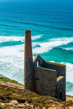 Old stone building with chimney of old tin mine on hillside with blue sea in the background; Cornwall County, England