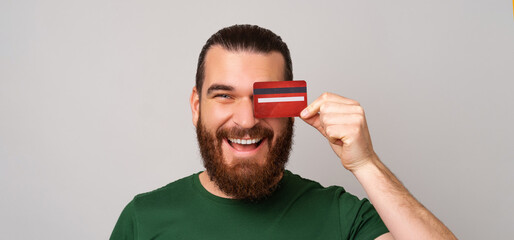 Panorama shot of an ecstatic bearded man covers one eye with a red credit card while smiling.