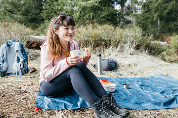 Young beautiful woman drinking hot tea or coffee from a thermos in the forest on a walk. Hiking