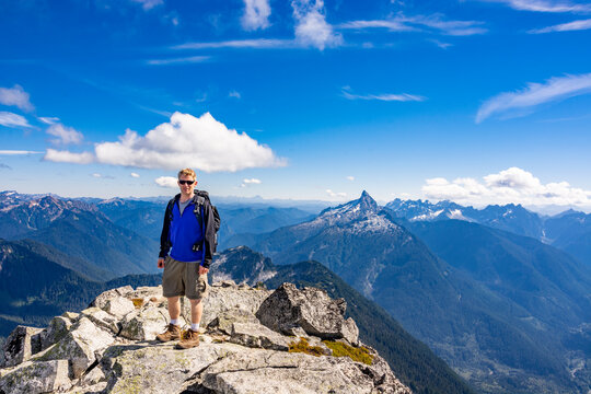 Adventurous athletic male hiker standing on top of a rugged mountain in the Pacific Northwest with jagged mountains in the background.