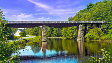 Fototapeta na wymiar Bridge casts a reflection into the river's calm water on a pleasant summer day in rural Nova Scotia, Canada.