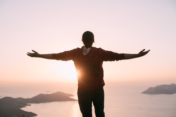 Fototapeta premium A young man meets the sunset, arms outstretched in a free gesture on a mountain by the sea.