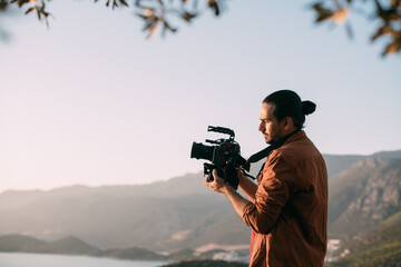 A young Caucasian man with a professional camera in his hands on a mountain by the sea. - obrazy, fototapety, plakaty