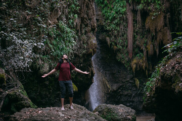 A young, joyful male tourist stands with his arms outstretched near a tropical waterfall among the rocks.